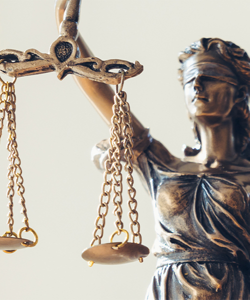 The image features the Statue of Liberty, a bronze sculpture representing justice. The statue is depicted blindfolded, symbolizing impartiality and the pursuit of truth. Her right hand holds a set of scales, which are divided into two pans by chains. The left pan appears heavier, indicating that the scale is currently biased in favor of the prosecution, and the right pan is lighter, suggesting the defendant's case. The statue stands against a plain, light-colored background, which emphasizes the symbol of justice itself. The image conveys a powerful message about the legal system's commitment to fairness and equal treatment under the law.